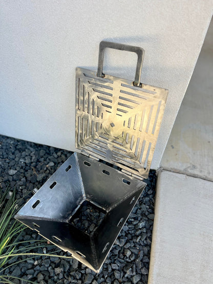 metal rocket stove on a forest floor with trees in the background camping and high heat cooking fireplace with grill top attachment to cook on top