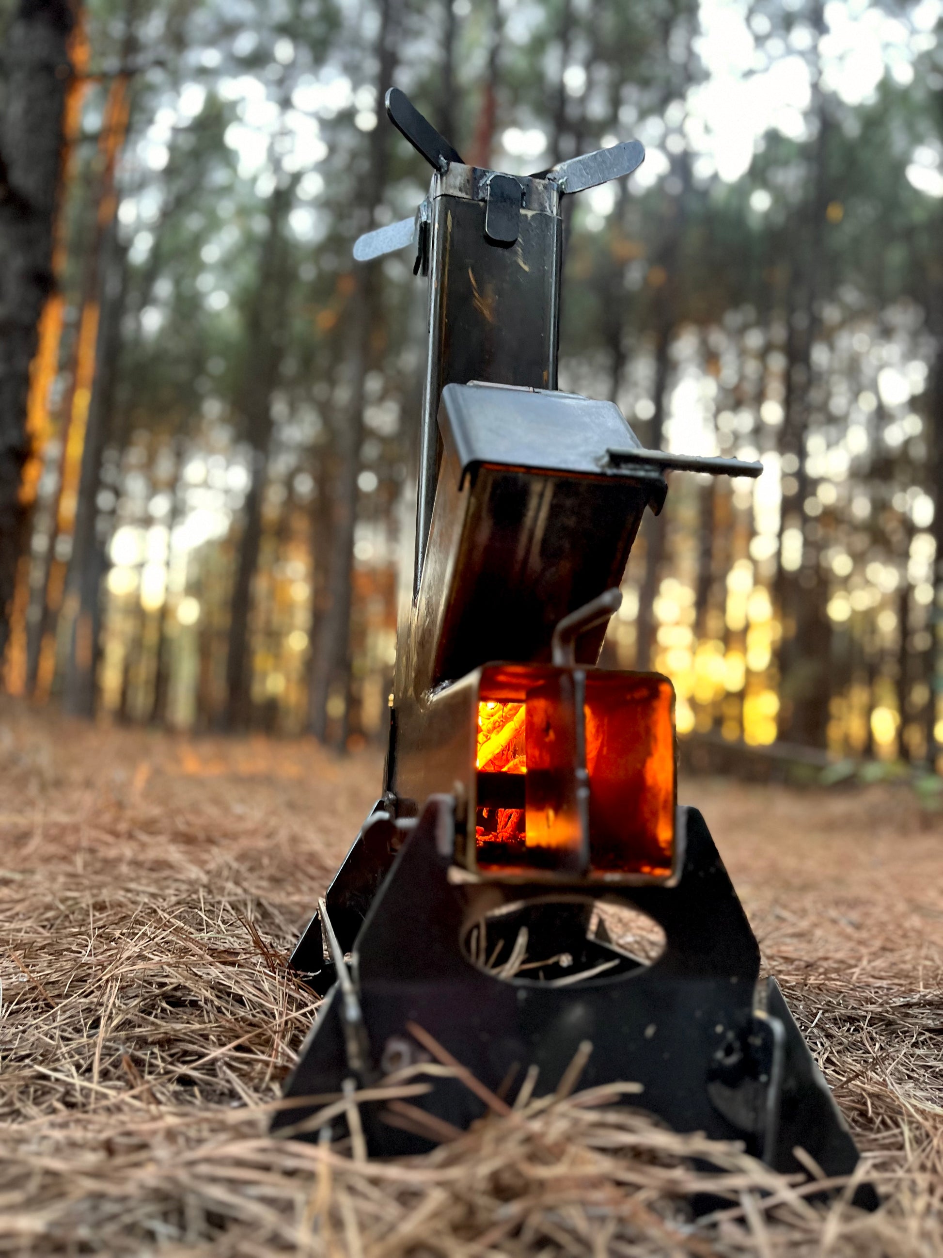 metal rocket stove on a forest floor with trees in the background camping and high heat cooking fireplace with flame and burn showing