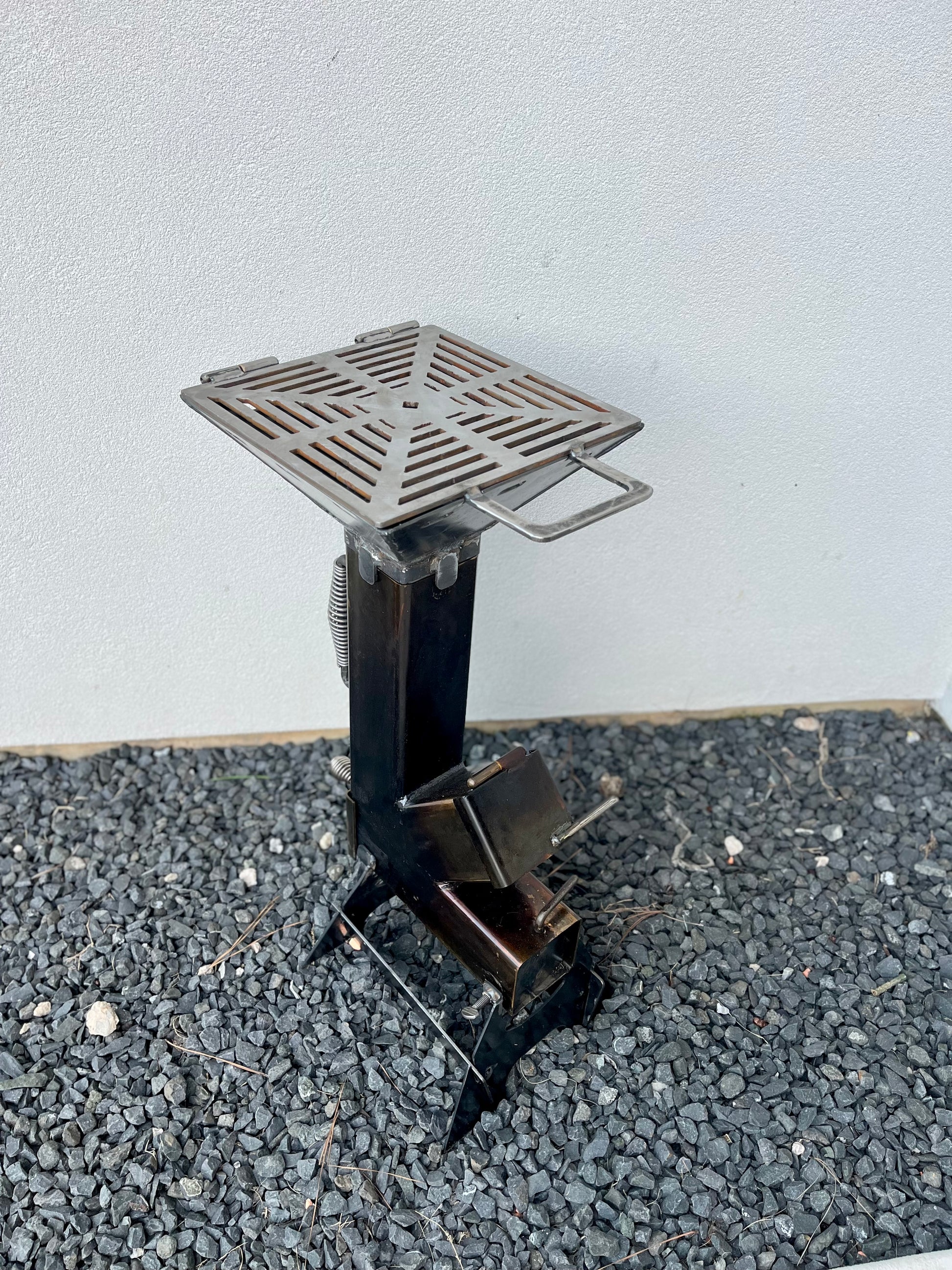 Small metal  grill top on rocket stove on a gray surface with a light gray background metal rocket stove on a forest floor with trees in the background camping and high heat cooking fireplace with grill top attachment to cook on top