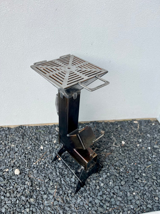 Small metal  grill top on rocket stove on a gray surface with a light gray background metal rocket stove on a forest floor with trees in the background camping and high heat cooking fireplace with grill top attachment to cook on top