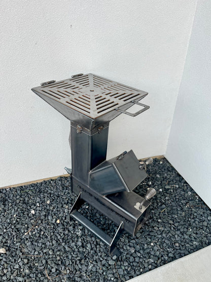 metal rocket stove on a forest floor with trees in the background camping and high heat cooking fireplace with grill top attachment to cook on top