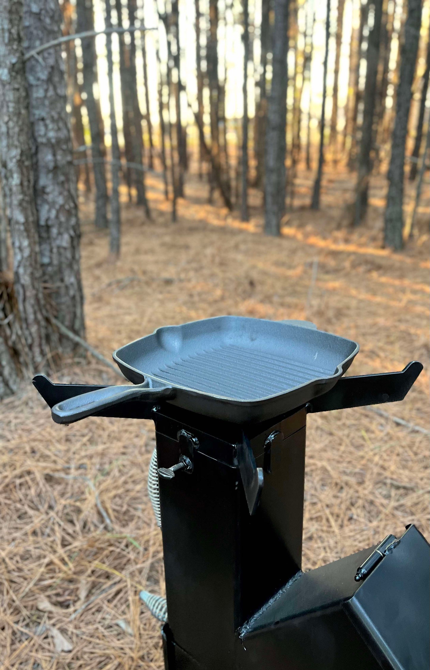 metal rocket stove on a forest floor with trees in the background camping and high heat cooking fireplace with cook cast iron skillet