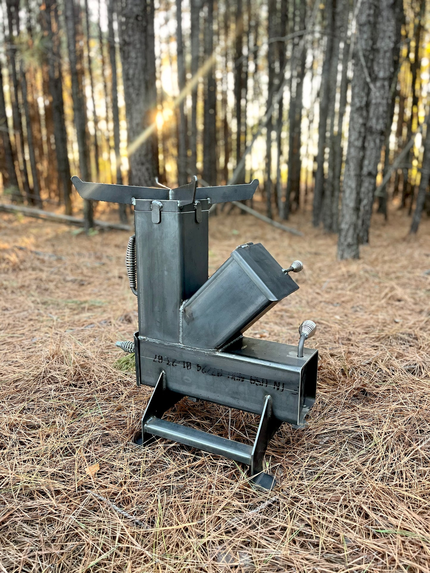 metal rocket stove on a forest floor with trees in the background camping and high heat cooking fireplace