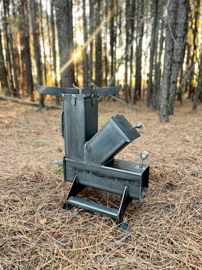 metal rocket stove on a forest floor with trees in the background camping and high heat cooking fireplace