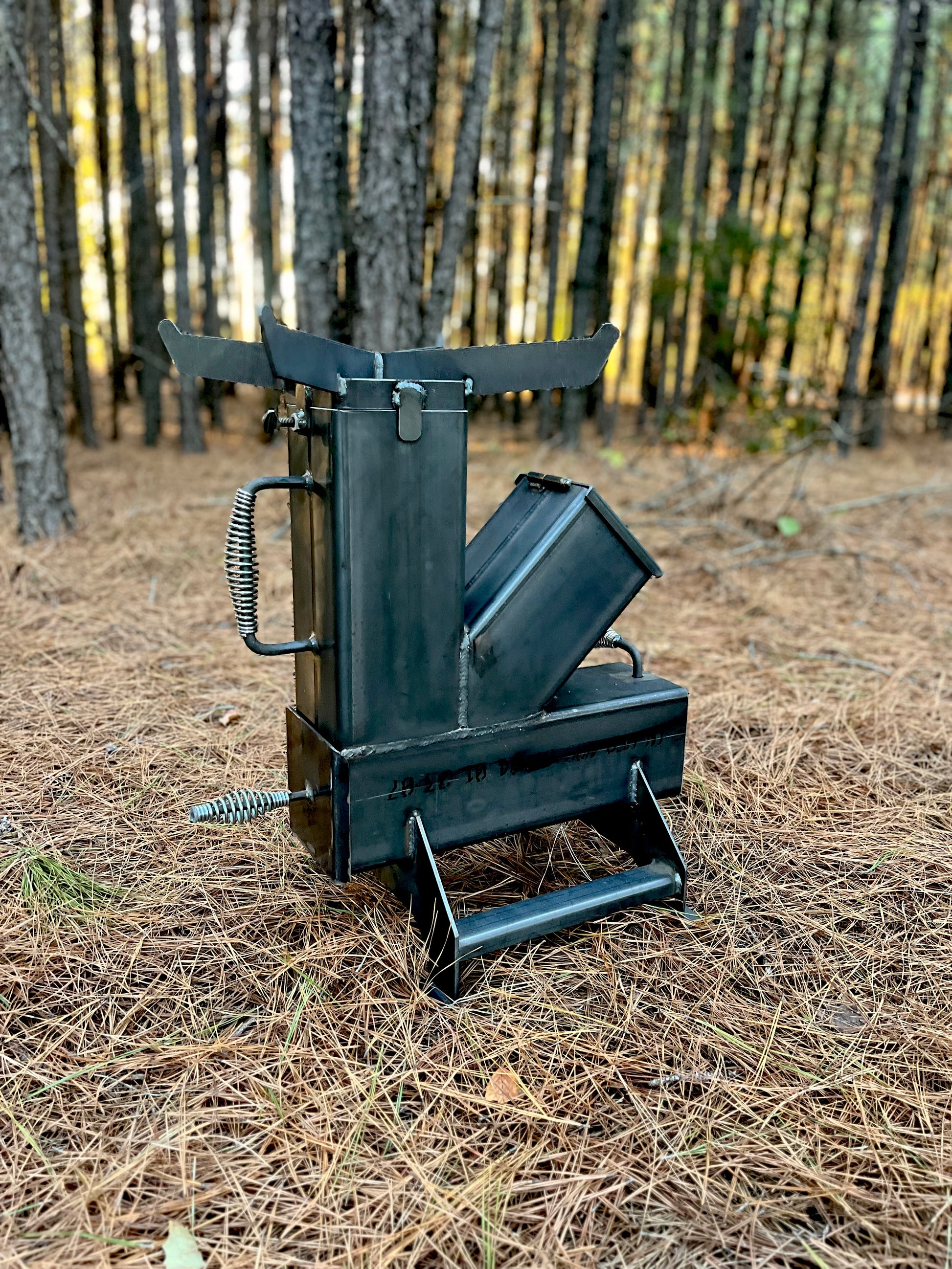 metal rocket stove on a forest floor with trees in the background camping and high heat cooking fireplace