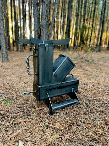 metal rocket stove on a forest floor with trees in the background camping and high heat cooking fireplace