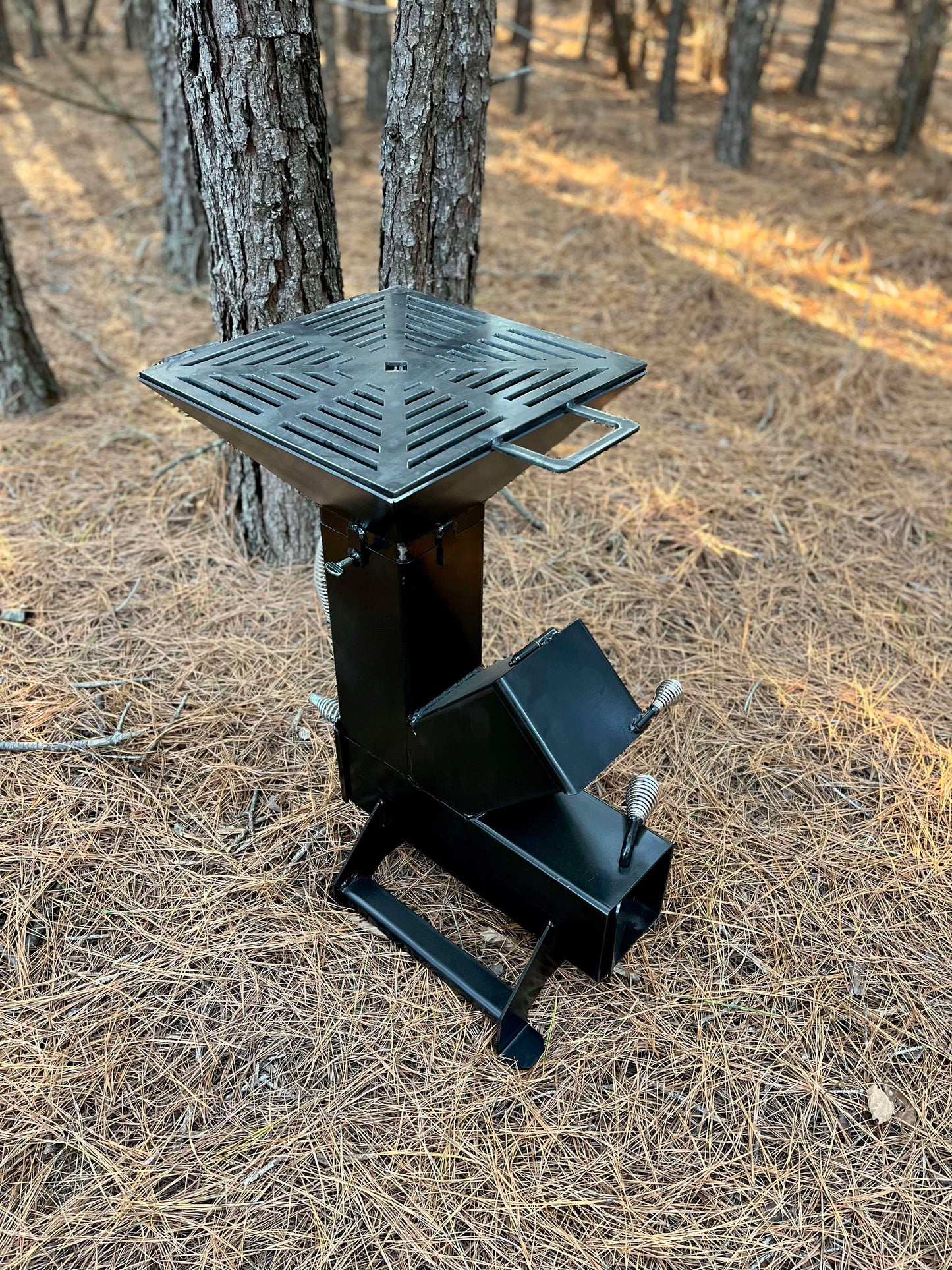 metal rocket stove on a forest floor with trees in the background camping and high heat cooking fireplace with grill top to cook on