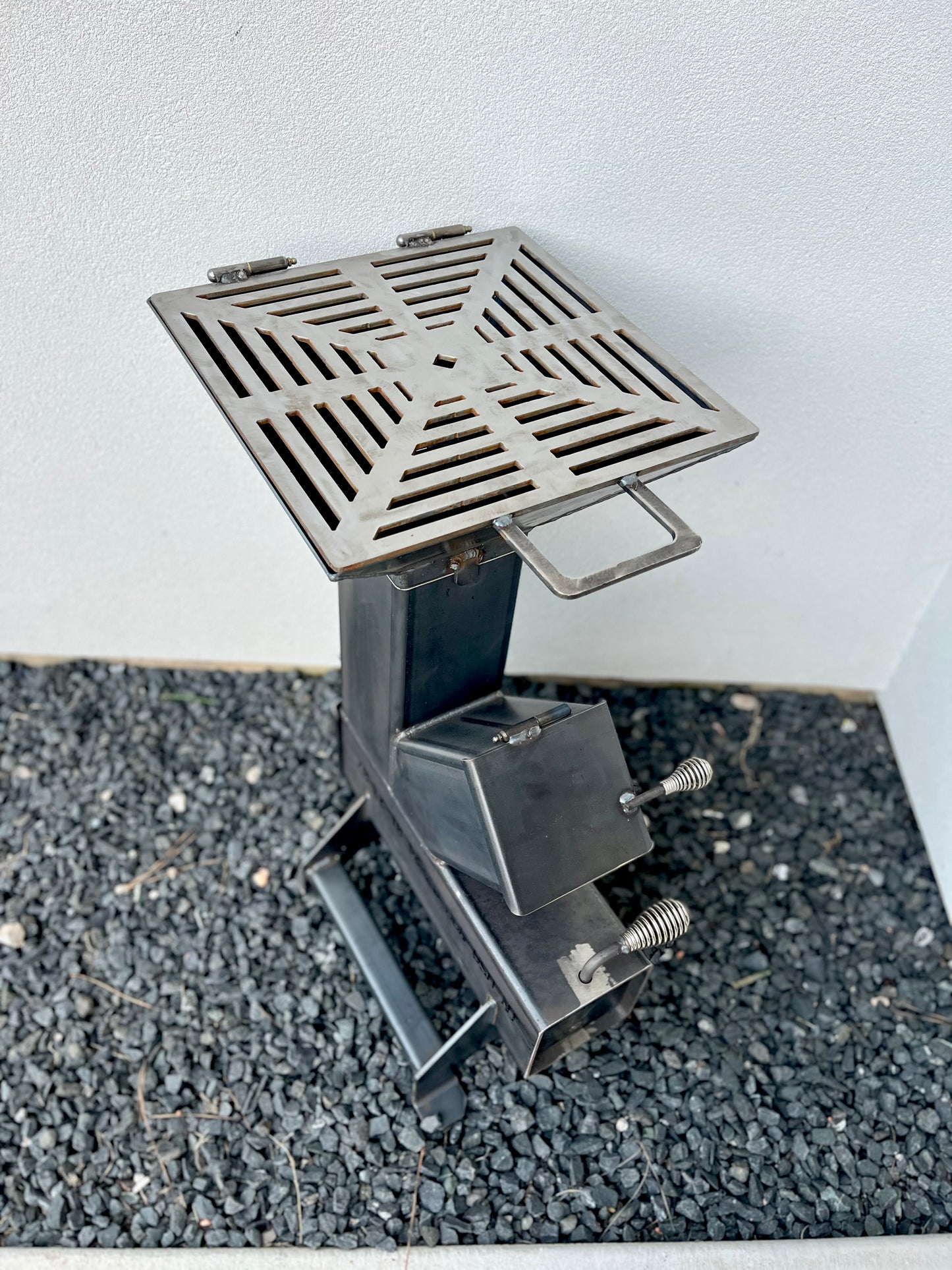 metal rocket stove on a forest floor with trees in the background camping and high heat cooking fireplace with grill top attachment to cook on