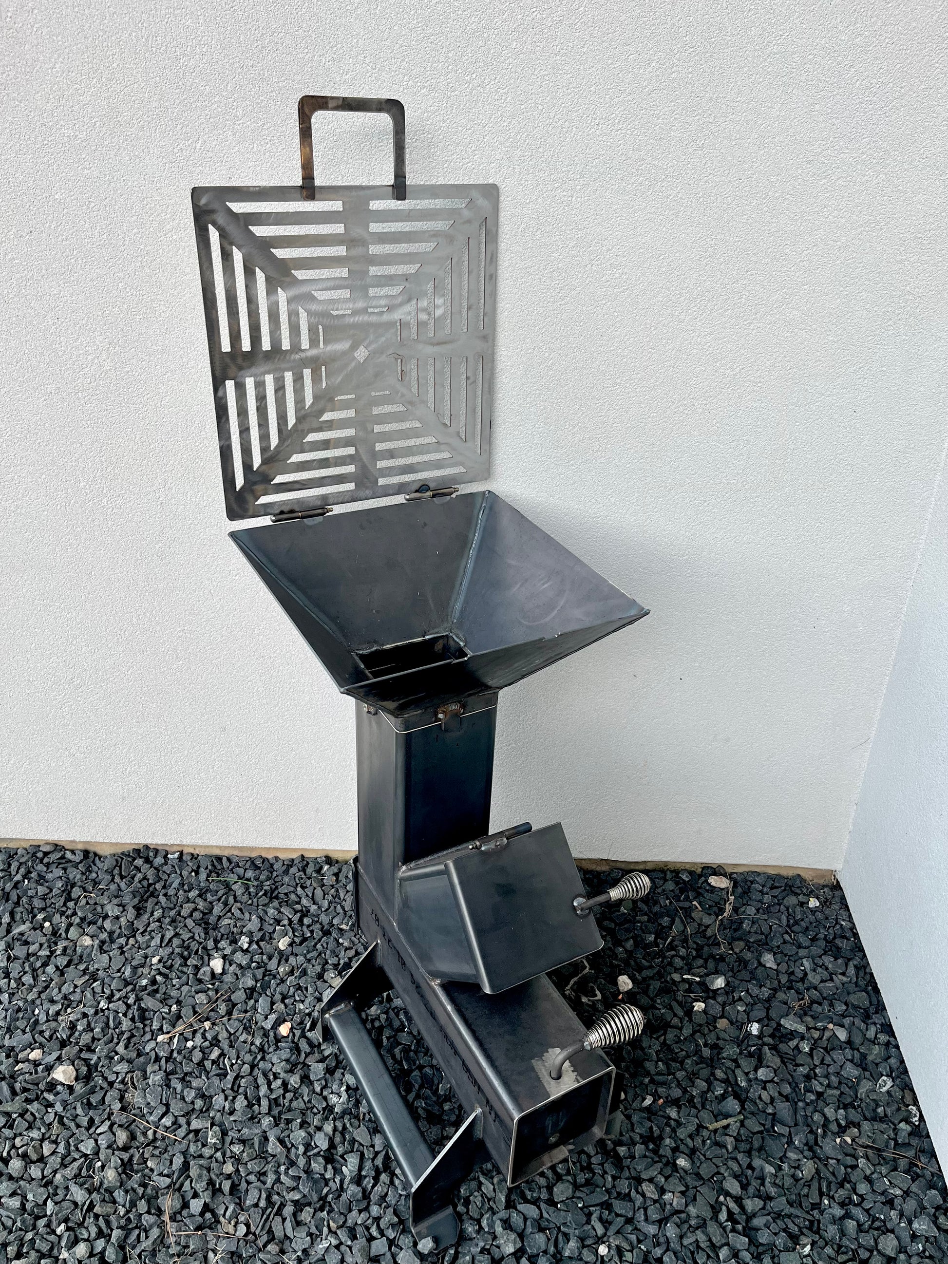 metal rocket stove on a forest floor with trees in the background camping and high heat cooking fireplace with grill top attachment to cook on top