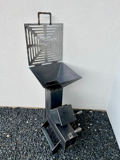 metal rocket stove on a forest floor with trees in the background camping and high heat cooking fireplace with grill top attachment to cook on top