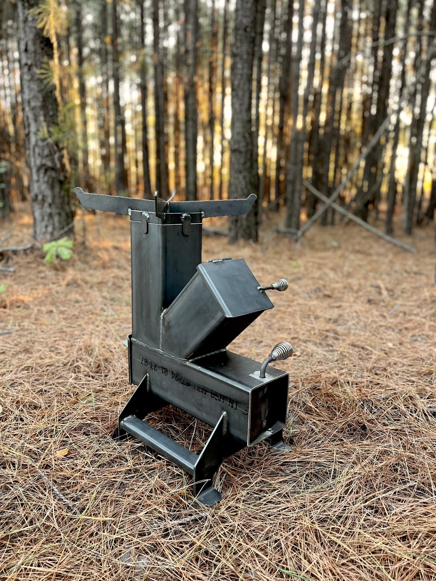 metal rocket stove on a forest floor with trees in the background camping and high heat cooking fireplace