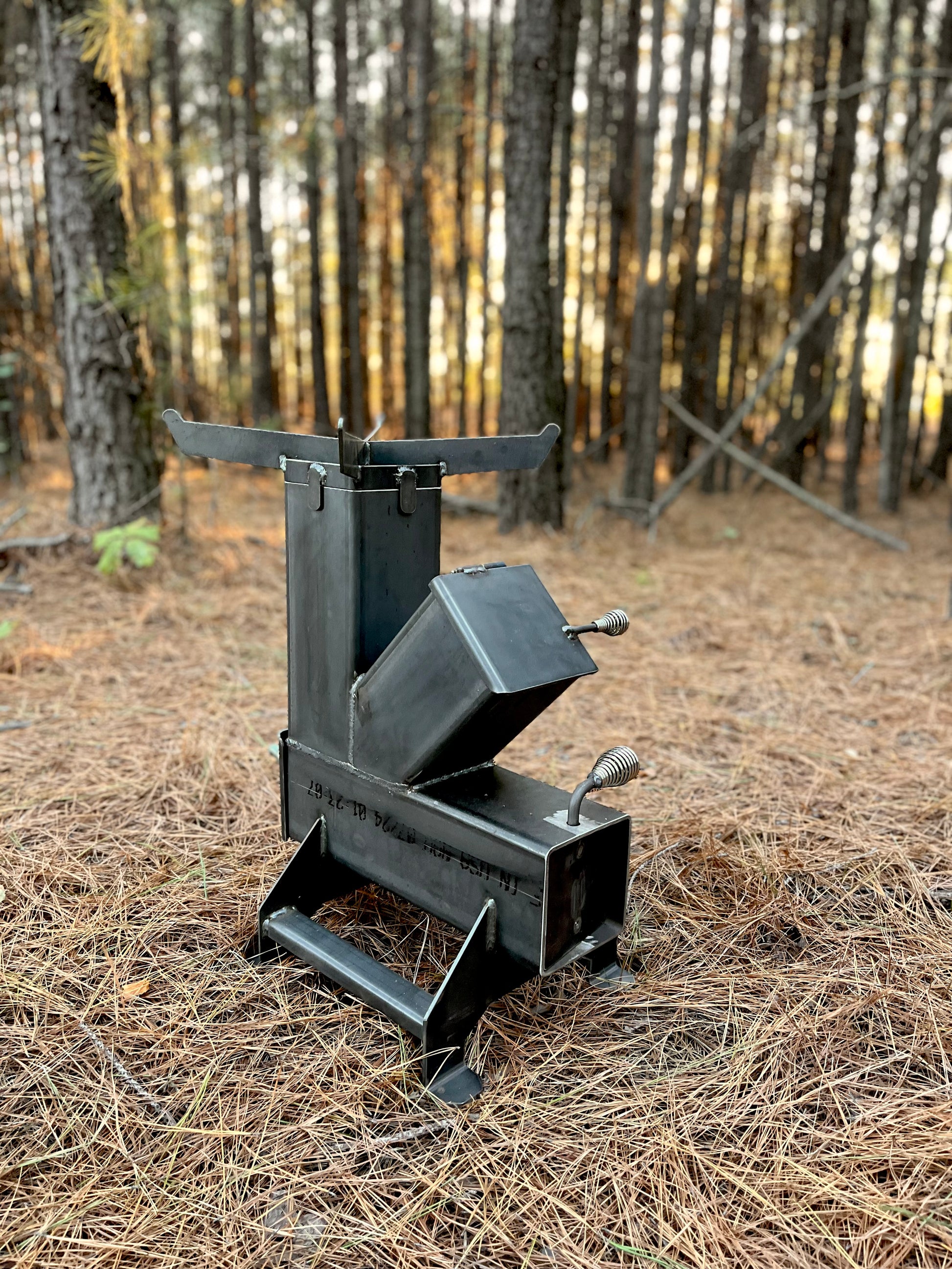 metal rocket stove on a forest floor with trees in the background camping and high heat cooking fireplace