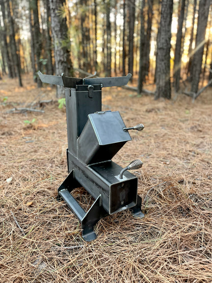 metal rocket stove on a forest floor with trees in the background camping and high heat cooking fireplace