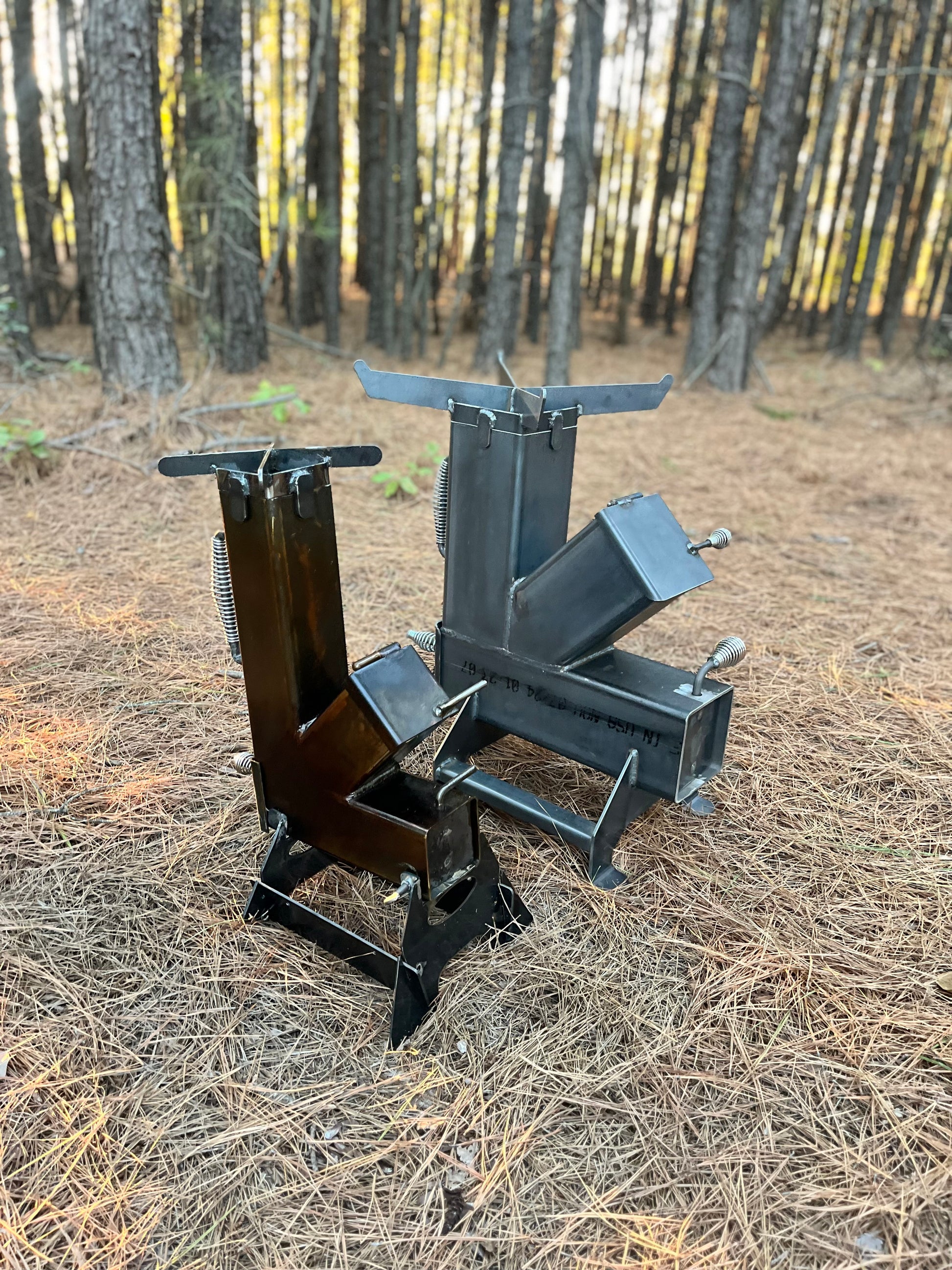 metal rocket stove on a forest floor with trees in the background camping and high heat cooking fireplace