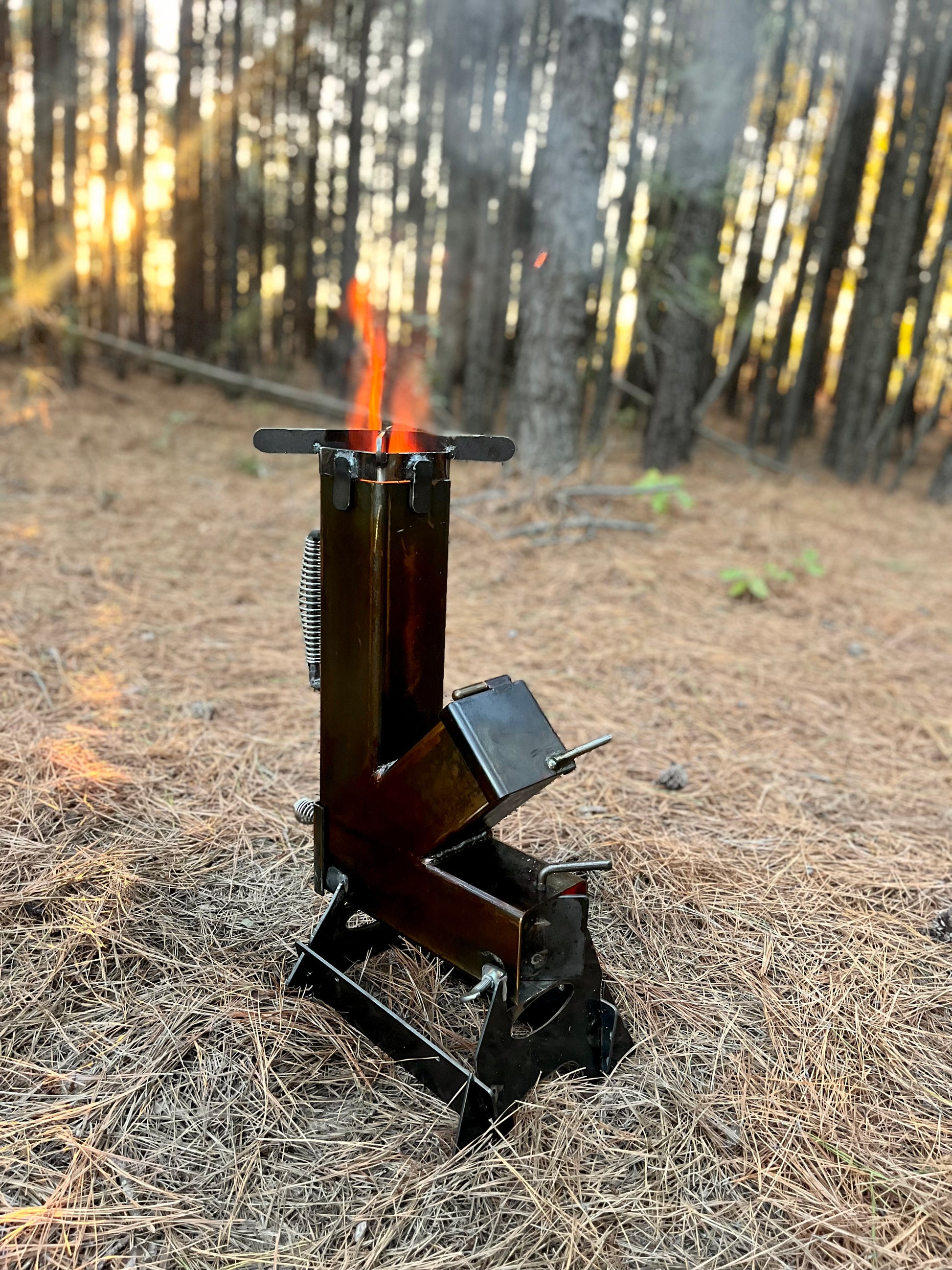 metal rocket stove on a forest floor with trees in the background camping and high heat cooking fireplace and lit fire
