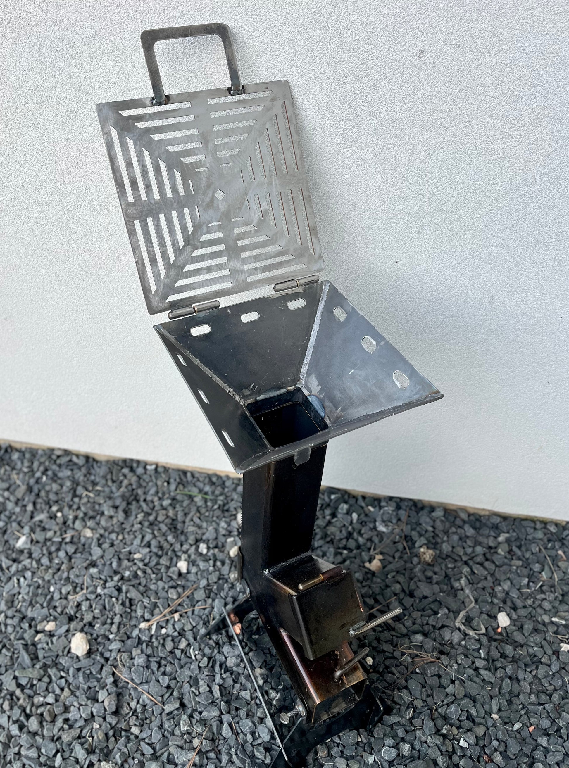 Metal grill on a gray surface rocket stove grill top attachment metal rocket stove on a forest floor with trees in the background camping and high heat cooking fireplace with grill top attachment to cook on top