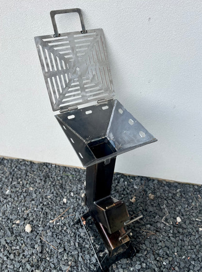 Metal grill on a gray surface rocket stove grill top attachment metal rocket stove on a forest floor with trees in the background camping and high heat cooking fireplace with grill top attachment to cook on top